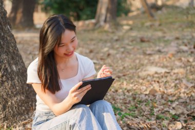 Genç Asyalı kadınlar, doğa parkındaki ağacın altında çimenlerde oturur ve iş bilgileri yazmak için dijital tablet kullanır ve seyahat yaşam tarzıyla rahatlarlar..