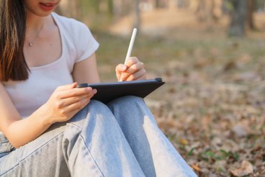 Genç Asyalı kadınlar dijital tablet üzerine iş verileri ve seyahat günlüğü yazarken doğa parkındaki çimlerin altında oturup dışarıda çalışmak ve seyahat yaşam tarzıyla rahatlamak..