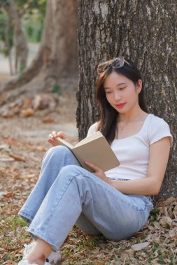 Genç Asyalı kadınlar çime yaslanıp, doğa parkında seyahat yaşam tarzıyla rahatlarken, eğlencesine edebiyat kitabı okurlar..