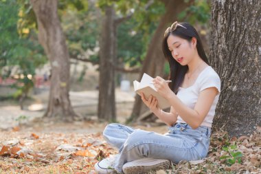 Genç Asyalı kadınlar, ağaçların altında çimlerin üzerinde oturup iş verileri ve seyahat süt ürünleri yazarken doğa parkındaki seyahat yaşam tarzlarıyla keyif çatıp, keyif çatıyorlar..