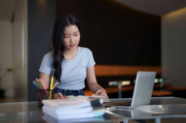 Asyalı genç kız öğrenci kitap okuyor ve ödev yaparken dijital dizüstü bilgisayarla ilgili ders seyrediyor ve kafede eğitim bilgisi öğreniyor..
