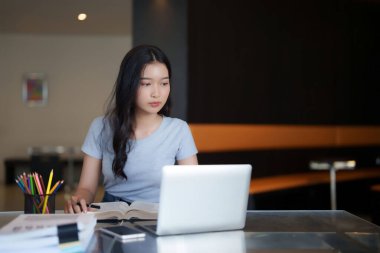 Asyalı genç bayan, internetten video izledikten ve kafede eğitim öğrenimi gördükten sonra ödev olarak dizüstü bilgisayardaki verileri araştırmak ve not almak için kitap okuyor..