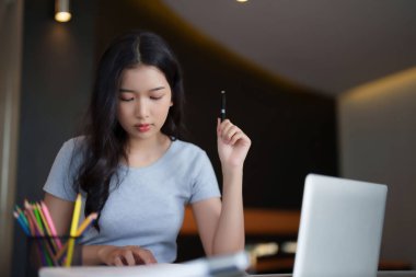 Asyalı genç bayan, ödev ödevi için not almak üzere kitap okuyor ve kafede eğitim bilgisi öğrenme ve öğrenme üzerine online dizüstü bilgisayar dersi izliyor..