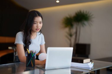 Asyalı genç kız öğrenci kitap okuyor ve internetten ders izledikten ve kafede eğitim öğrenimi gördükten sonra ödev yapmak için dizüstü bilgisayarda veri araştırması yapıyor..