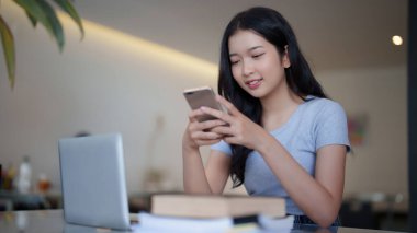 Asyalı genç bayan, bilgisayardaki videoyu izledikten ve kafede eğitim bilgisi öğrendikten sonra arkadaşlarıyla sohbet etmek ve sosyal medyada sörf yapmak için akıllı telefon kullanıyor..