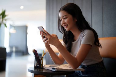 Asyalı genç bayan, kafeteryadaki eğitim bilgisi hakkında bilgi edinmek için okuduktan sonra arkadaşlarıyla sohbet etmek ve sosyal medyada sörf yapmak için akıllı telefon kullanıyor..