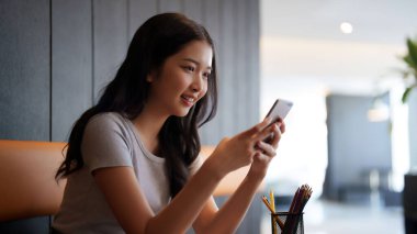 Asyalı genç bayan, kafeteryadaki eğitim bilgisi hakkında bilgi edinmek için okuduktan sonra arkadaşlarıyla sohbet etmek ve sosyal medyada sörf yapmak için akıllı telefon kullanıyor..