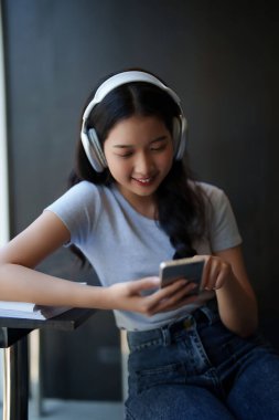 Asyalı genç bayan arkadaşlarıyla sohbet etmek için akıllı telefon kullanıyor ve kafede sınav hazırlamak için eğitim bilgisi öğrendikten sonra kulaklık takıp mutlulukla müzik dinliyor..