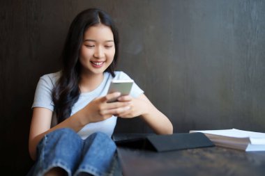 Asyalı genç bayan sosyal medyada sörf yapmak için akıllı telefonu kullanıyor ve kafeteryada eğitim hakkında bilgi edinmek için kağıt okuduktan sonra arkadaşlarıyla mutlulukla sohbet ediyor..