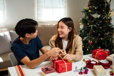 Genç bir adam ve kadın bir masada birlikte oturuyorlar, hediyeleri hoş, şenlikli bir ortamda paketliyorlar. Arka planda süslü bir Noel ağacı görünüyor