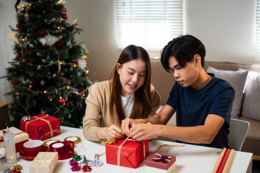 Genç bir adam ve kadın bir masada birlikte oturuyorlar, hediyeleri hoş, şenlikli bir ortamda paketliyorlar. Arka planda süslü bir Noel ağacı görünüyor