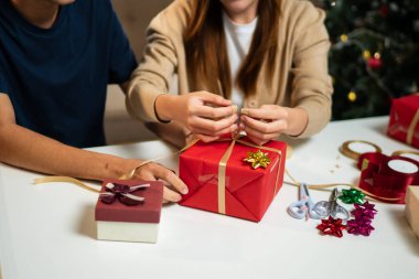 Genç bir adam ve kadın bir masada birlikte oturuyorlar, hediyeleri hoş, şenlikli bir ortamda paketliyorlar. Arka planda süslü bir Noel ağacı görünüyor