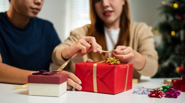 Genç bir adam ve kadın bir masada birlikte oturuyorlar, hediyeleri hoş, şenlikli bir ortamda paketliyorlar. Arka planda süslü bir Noel ağacı görünüyor