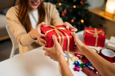 Genç bir adam ve kadın bir masada oturuyor, bayram ortamında birbirlerine bir hediye veriyorlar. Arka planda süslü bir Noel ağacı