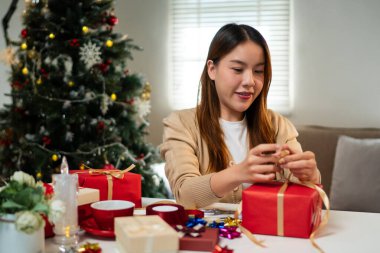 Genç bir kadın, bayram süslemeleriyle çevrili, odaklanmış ve neşeli bir masada hediyeler paketliyor. Arka planda bir Noel ağacı 