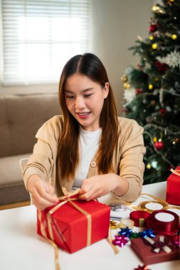 Genç bir kadın, bayram süslemeleriyle çevrili, odaklanmış ve neşeli bir masada hediyeler paketliyor. Arka planda bir Noel ağacı 