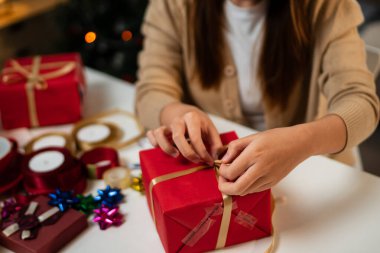 Genç bir kadın, bayram süslemeleriyle çevrili, odaklanmış ve neşeli bir masada hediyeler paketliyor. Arka planda bir Noel ağacı 