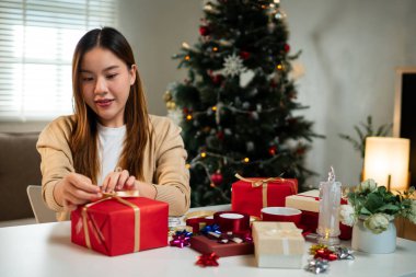 Genç bir kadın, bayram süslemeleriyle çevrili, odaklanmış ve neşeli bir masada hediyeler paketliyor. Arka planda bir Noel ağacı 