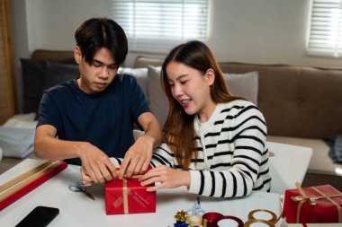 Genç bir adam ve kadın bir masada oturuyor, mutlu bir şekilde hediyeleri paketliyorlar. Tatil süsleriyle çevrili neşeli bir anı paylaşıyorlar.