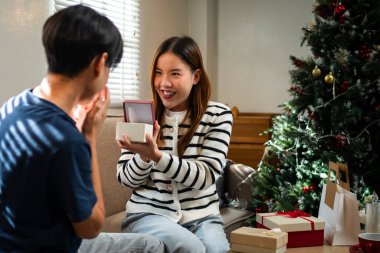 Genç bir adam ve bir kadın Noel ağacının yanında oturmuş neşe dolu bir anı paylaşıyorlar. Kadın elinde bir hediyeyle heyecanlı bir şekilde gülümsüyor..