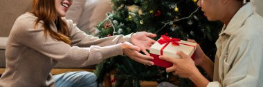 İki el güzel paketlenmiş kırmızı ve beyaz bir hediye kutusunu kırmızı bir fiyonkla değiş tokuş eder, süslü bir Noel ağacının arka planına kurulur, sıcak bir tatil anı yaratır..