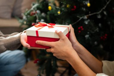 İki el güzel paketlenmiş kırmızı ve beyaz bir hediye kutusunu kırmızı bir fiyonkla değiş tokuş eder, süslü bir Noel ağacının arka planına kurulur, sıcak bir tatil anı yaratır..