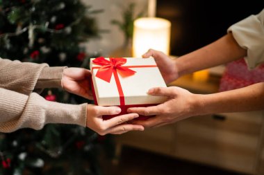 İki el güzel paketlenmiş kırmızı ve beyaz bir hediye kutusunu kırmızı bir fiyonkla değiş tokuş eder, süslü bir Noel ağacının arka planına kurulur, sıcak bir tatil anı yaratır..
