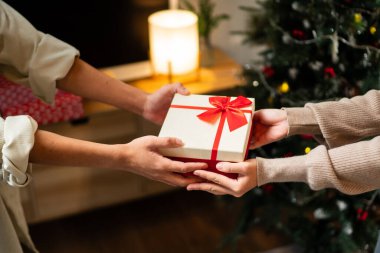 İki el güzel paketlenmiş kırmızı ve beyaz bir hediye kutusunu kırmızı bir fiyonkla değiş tokuş eder, süslü bir Noel ağacının arka planına kurulur, sıcak bir tatil anı yaratır..