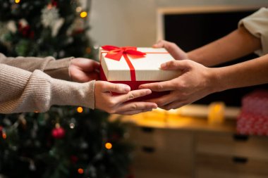 İki el güzel paketlenmiş kırmızı ve beyaz bir hediye kutusunu kırmızı bir fiyonkla değiş tokuş eder, süslü bir Noel ağacının arka planına kurulur, sıcak bir tatil anı yaratır..