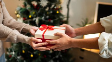 İki el güzel paketlenmiş kırmızı ve beyaz bir hediye kutusunu kırmızı bir fiyonkla değiş tokuş eder, süslü bir Noel ağacının arka planına kurulur, sıcak bir tatil anı yaratır..