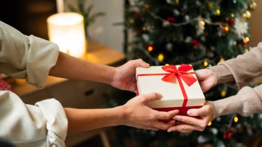 İki el güzel paketlenmiş kırmızı ve beyaz bir hediye kutusunu kırmızı bir fiyonkla değiş tokuş eder, süslü bir Noel ağacının arka planına kurulur, sıcak bir tatil anı yaratır..