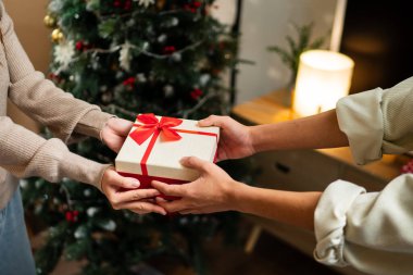 İki el güzel paketlenmiş kırmızı ve beyaz bir hediye kutusunu kırmızı bir fiyonkla değiş tokuş eder, süslü bir Noel ağacının arka planına kurulur, sıcak bir tatil anı yaratır..