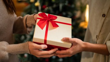 İki el güzel paketlenmiş kırmızı ve beyaz bir hediye kutusunu kırmızı bir fiyonkla değiş tokuş eder, süslü bir Noel ağacının arka planına kurulur, sıcak bir tatil anı yaratır..