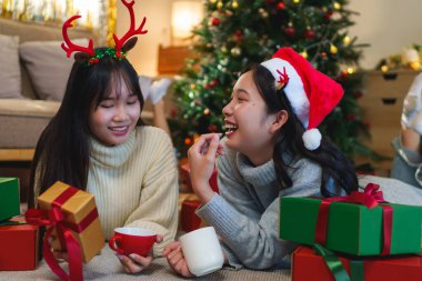  İki arkadaş, süslü bir Noel ağacının yanında yerde uzanmış, sıcak içeceklerin tadını çıkarıyor ve gülüyor, sıcacık bir tatil ortamında paketlenmiş hediyelerle çevrili..