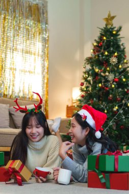  İki arkadaş, süslü bir Noel ağacının yanında yerde uzanmış, sıcak içeceklerin tadını çıkarıyor ve gülüyor, sıcacık bir tatil ortamında paketlenmiş hediyelerle çevrili..