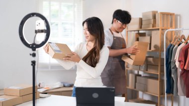  Bir kadın elinde bir kutu tutarken heyecanla yaşıyor, ürünlerini raflarla ve paketlerle dolu bir çalışma alanında halka ışığı ve dizüstü bilgisayarla sergiliyor..