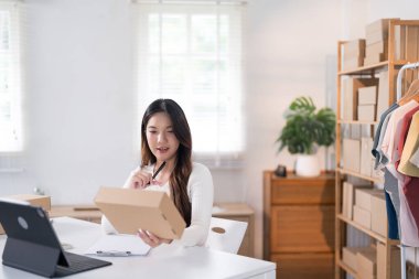 Bir kadın elinde bir kutu tutarken heyecanla yaşıyor, ürünlerini raflarla ve paketlerle dolu bir çalışma alanında halka ışığı ve dizüstü bilgisayarla sergiliyor..
