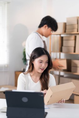 Bir kadın elinde bir kutu tutarken heyecanla yaşıyor, ürünlerini raflarla ve paketlerle dolu bir çalışma alanında halka ışığı ve dizüstü bilgisayarla sergiliyor..