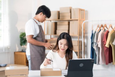 Bir kadın elinde bir kutu tutarken heyecanla yaşıyor, ürünlerini raflarla ve paketlerle dolu bir çalışma alanında halka ışığı ve dizüstü bilgisayarla sergiliyor..