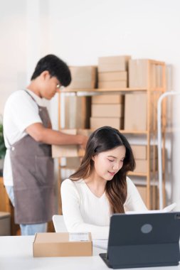 Bir kadın elinde bir kutu tutarken heyecanla yaşıyor, ürünlerini raflarla ve paketlerle dolu bir çalışma alanında halka ışığı ve dizüstü bilgisayarla sergiliyor..