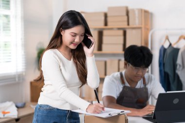 Bir kadın, teslimat kutuları ve giysi raflarıyla dolu bir çalışma alanında dizüstü bilgisayarında çalışan bir adamın yanında duran bir paketle telefonda konuşuyor..