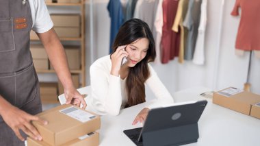 Çalışma odasındaki bir dizüstü bilgisayarda çalışan bir kadın, kutularla ve elbise raflarıyla çevriliyken telefonla konuşur..