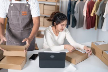 Bir kadın nakliye için bir paket hazırlarken, diğeri de kutularla ve elbise raflarıyla dolu bir çalışma alanında etiketi tarıyor..