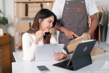 Bir erkek, bir kadın elbise askısı ve nakliye malzemeleriyle telefonuna bakarken bir kutu hazırlar..