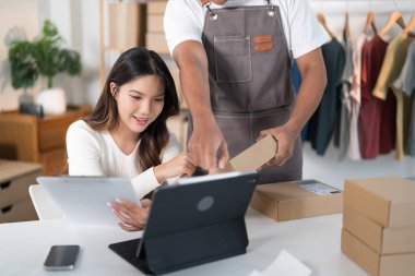 Bir erkek, bir kadın elbise askısı ve nakliye malzemeleriyle telefonuna bakarken bir kutu hazırlar..