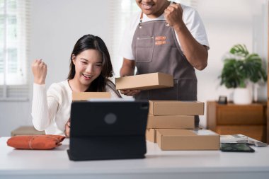 Neşeli bir çift online satışlardaki başarılarını kutluyor, kargo kutuları ve tabletlerle dolu bir çalışma alanında dururken ellerinde heyecanla paketler tutuyorlar..