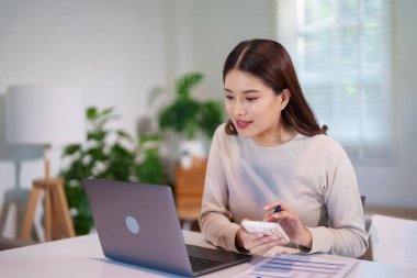 Bir kadın akıllı telefonu ve laptopuyla harcamaları hesaplıyor, finansal planlama ve işe odaklanıyor..