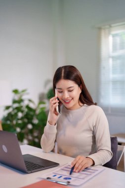 Bir kadın birden fazla iş yapıyor, bilgisayarını kullanırken telefonla konuşuyor ve masadaki belgeleri inceliyor..