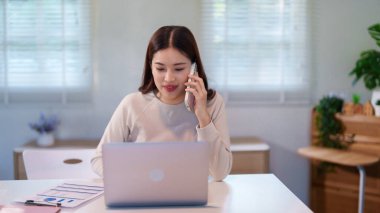 Bir kadın birden fazla iş yapıyor, bilgisayarını kullanırken telefonla konuşuyor ve masadaki belgeleri inceliyor..