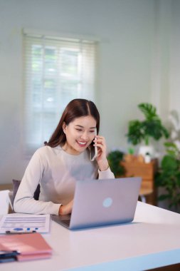 Bir kadın birden fazla iş yapıyor, bilgisayarını kullanırken telefonla konuşuyor ve masadaki belgeleri inceliyor..